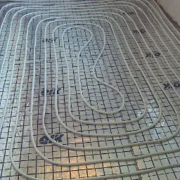 Białe rury ogrzewania podłogowego ułożone spiralnie na srebrnej macie z czarną siatką, widoczne fragmenty ściany w tle.